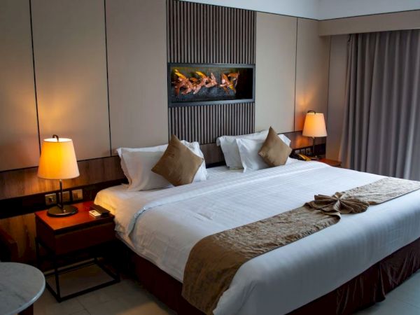A cozy hotel room with a large bed, decorative pillows, bedside lamps, and a wall-mounted fireplace, displaying a neat and inviting space.