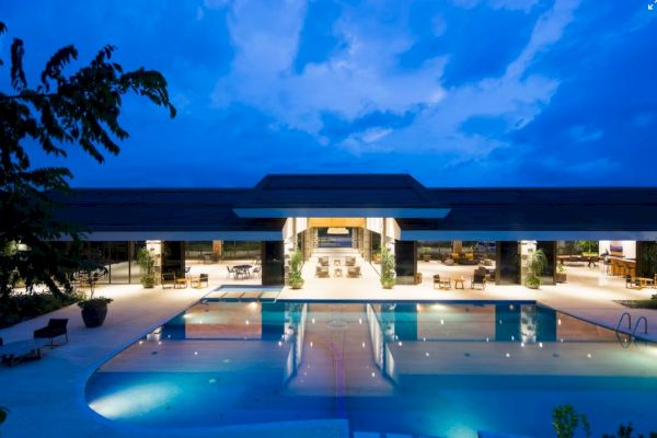 The image shows a modern building with a large, illuminated swimming pool in the foreground, surrounded by lounge chairs and trees at dusk.