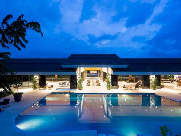 The image shows a modern building with a large, illuminated swimming pool in the foreground, surrounded by lounge chairs and trees at dusk.