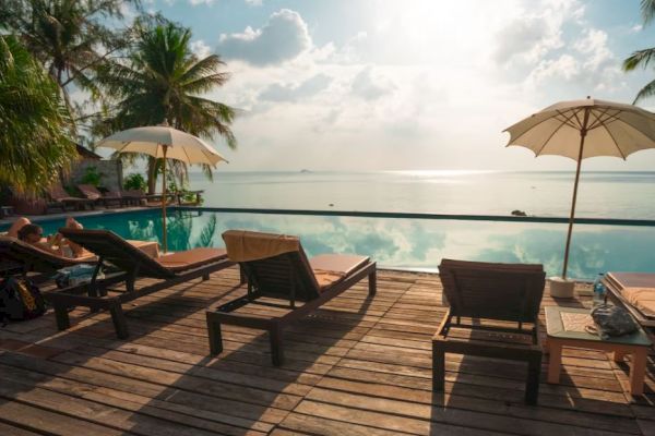 This image shows a serene poolside setting with lounge chairs, umbrellas, and a view of palm trees and a calm ocean under a partly cloudy sky.