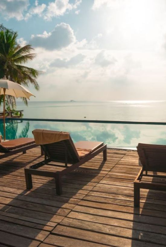 A serene poolside scene with lounge chairs, umbrellas, palm trees, and a reflection of clouds on the water, overlooking a calm ocean.