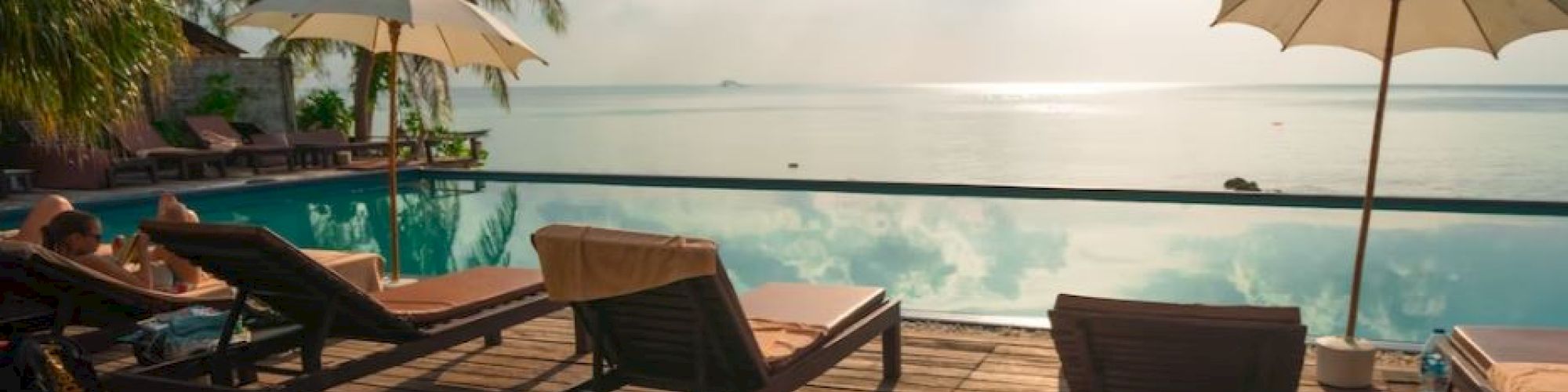 A tranquil poolside scene with lounge chairs and umbrellas overlooks a serene ocean under a partly cloudy sky, surrounded by palm trees.