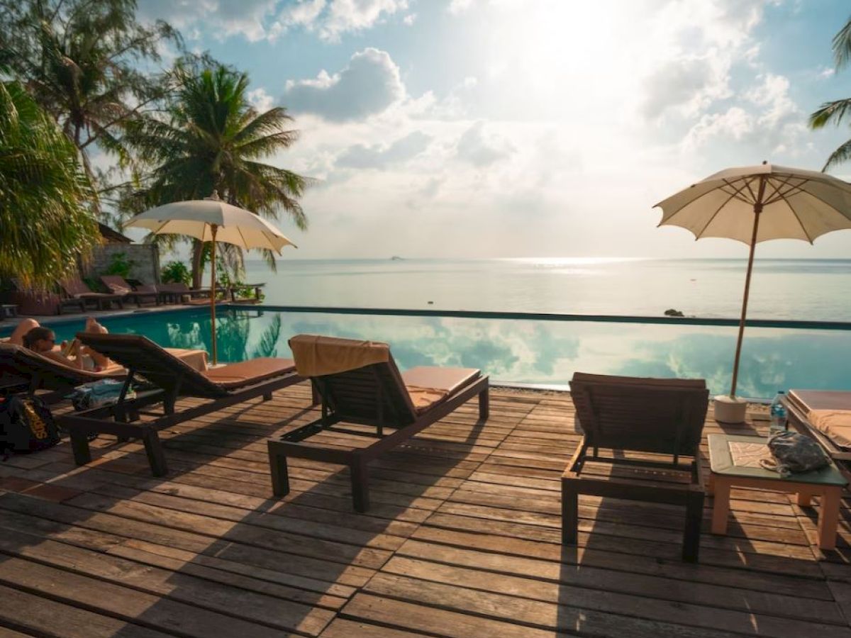 A serene tropical poolside scene with lounge chairs and umbrellas overlooking a calm, reflective pool and a beautiful seascape in the background.