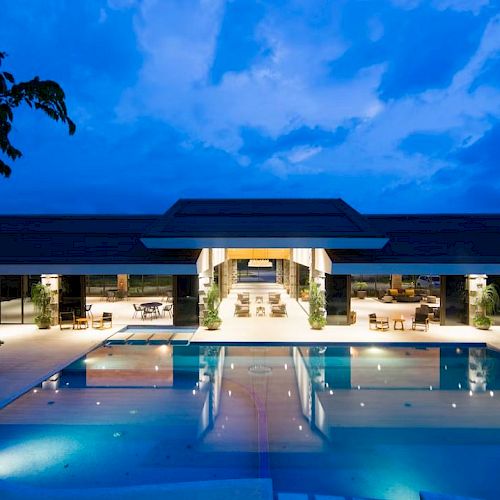A luxurious outdoor swimming pool area with lounge chairs and tables, illuminated under the evening sky and surrounded by open pavilion structures.