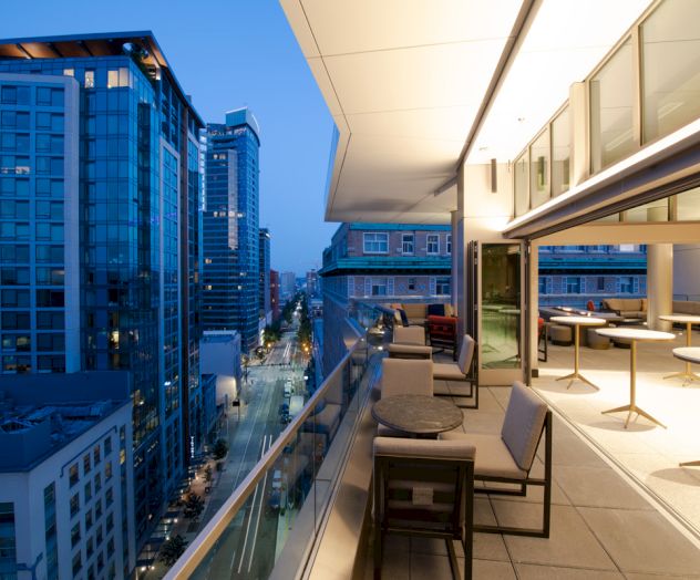 The image shows a modern cityscape with tall buildings viewed from a high balcony that has tables, chairs, and an open indoor-outdoor space.