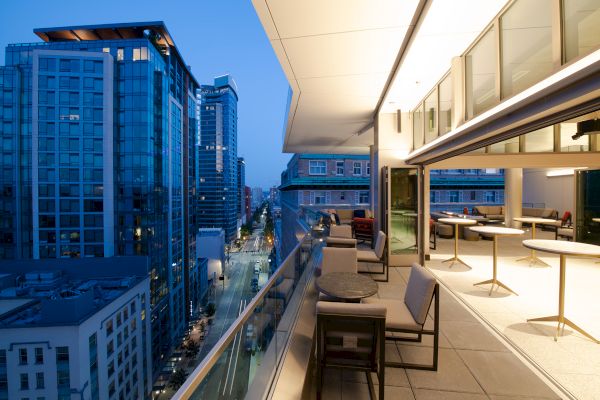 The image shows a furnished terrace overlooking a cityscape with tall, modern buildings at dusk. The terrace has seating and tables.