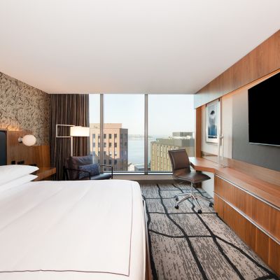 A modern hotel room with a large bed, a wall-mounted TV, a desk with a chair, and a large window offering a view of buildings and water outside the window.
