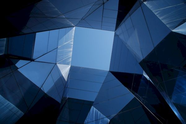The image shows a geometric arrangement of reflective glass panels creating a tunnel-like structure with an open sky visible at the top center.