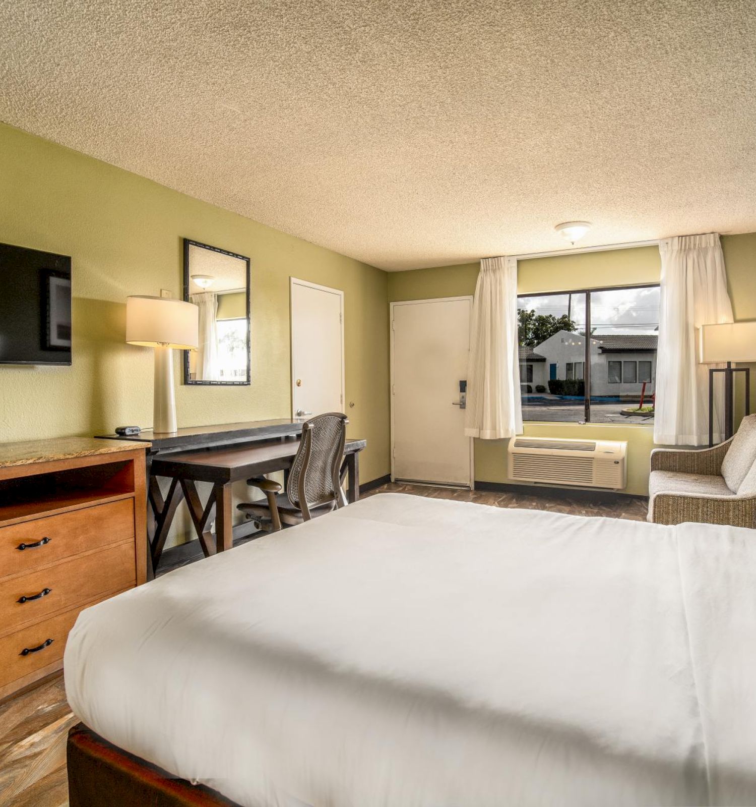 This image shows a hotel room with a bed, a TV on a dresser, a desk with a chair, and a couch. The room has green walls and a window with curtains.