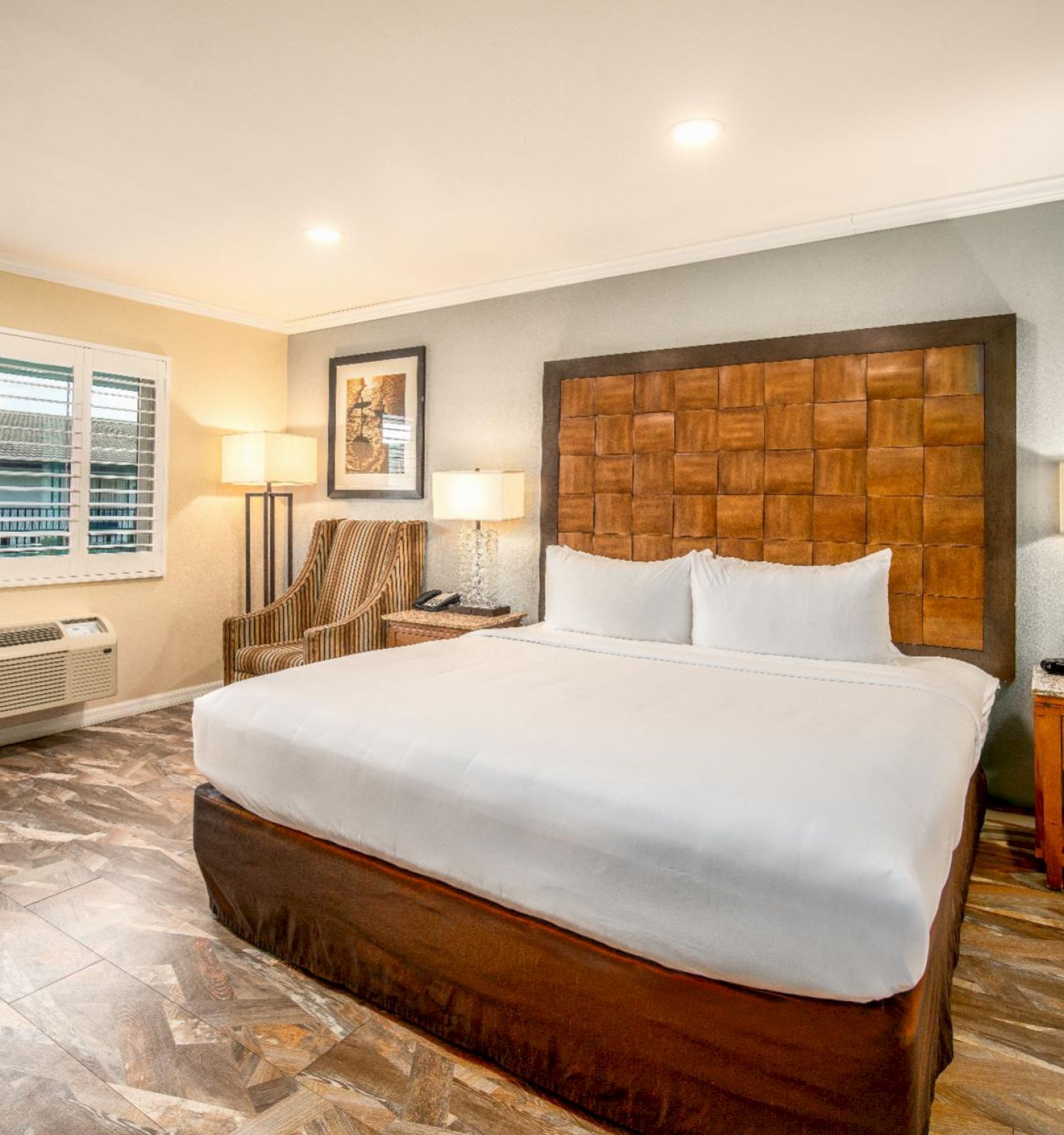 A hotel room with a king bed, nightstands, lamps, an armchair, a window with shutters, and tiled flooring is shown in the image.