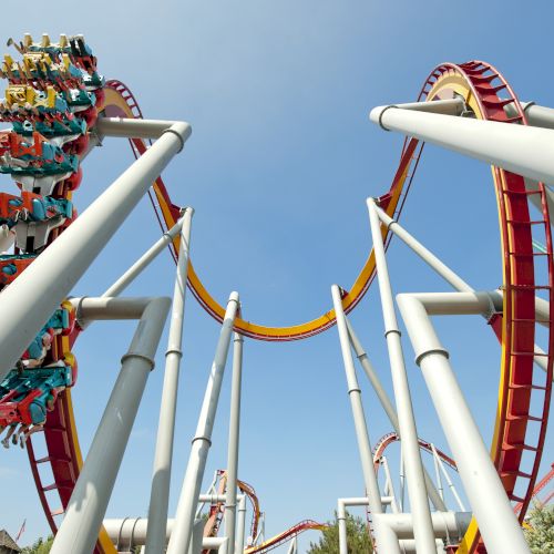 This image shows a roller coaster with loops and people riding in it, suspended upside down, against a clear blue sky with supporting structures visible.