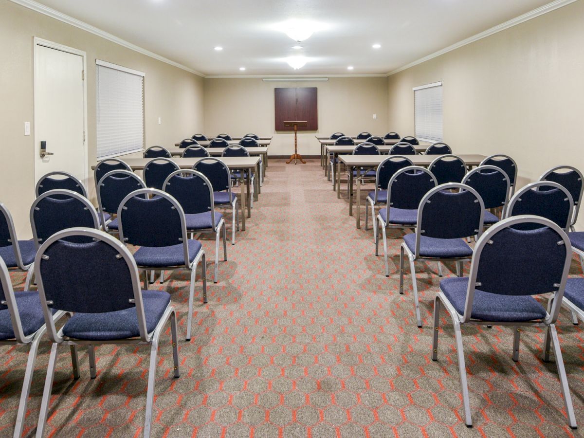 The image shows an empty conference room set up with rows of blue chairs facing a lectern at the front. The room has beige walls and carpet.