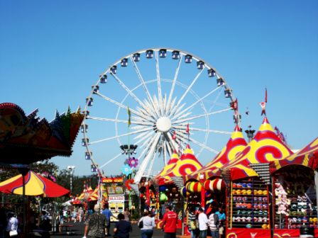 The image shows a carnival with a large Ferris wheel, colorful tents, game booths, and people walking around under a clear blue sky.