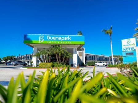 This image shows the exterior of Buena Park Hotel & Suites, featuring a sign and the building entrance, surrounded by palm trees and landscaped greenery.