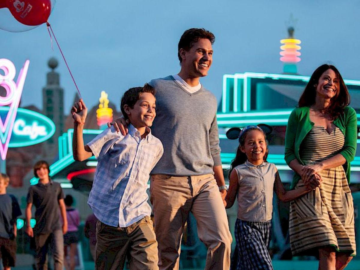 A family of four walks together at an amusement park; the boy holds a balloon, and neon lights illuminate the background.