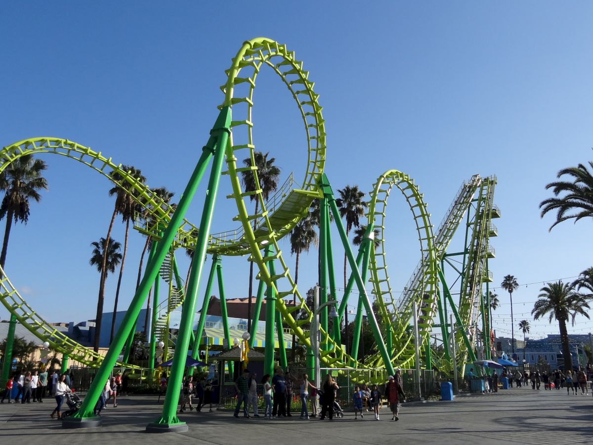 An amusement park scene shows a green roller coaster with loops and twists, palm trees in the background, and a clear blue sky.