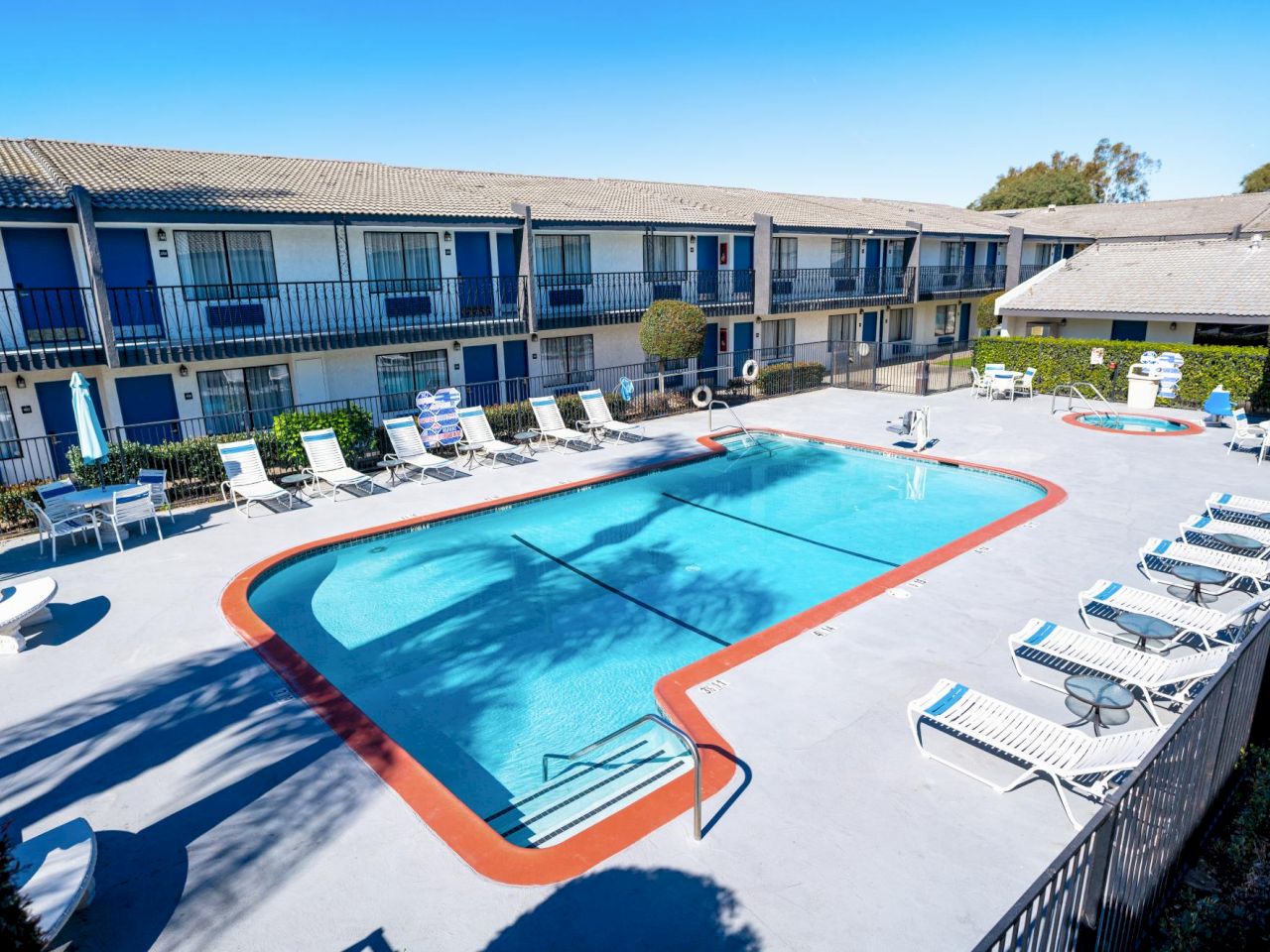 Aerial view of a hotel pool area with a kidney-shaped pool, lounge chairs, and a two-story building in the background.