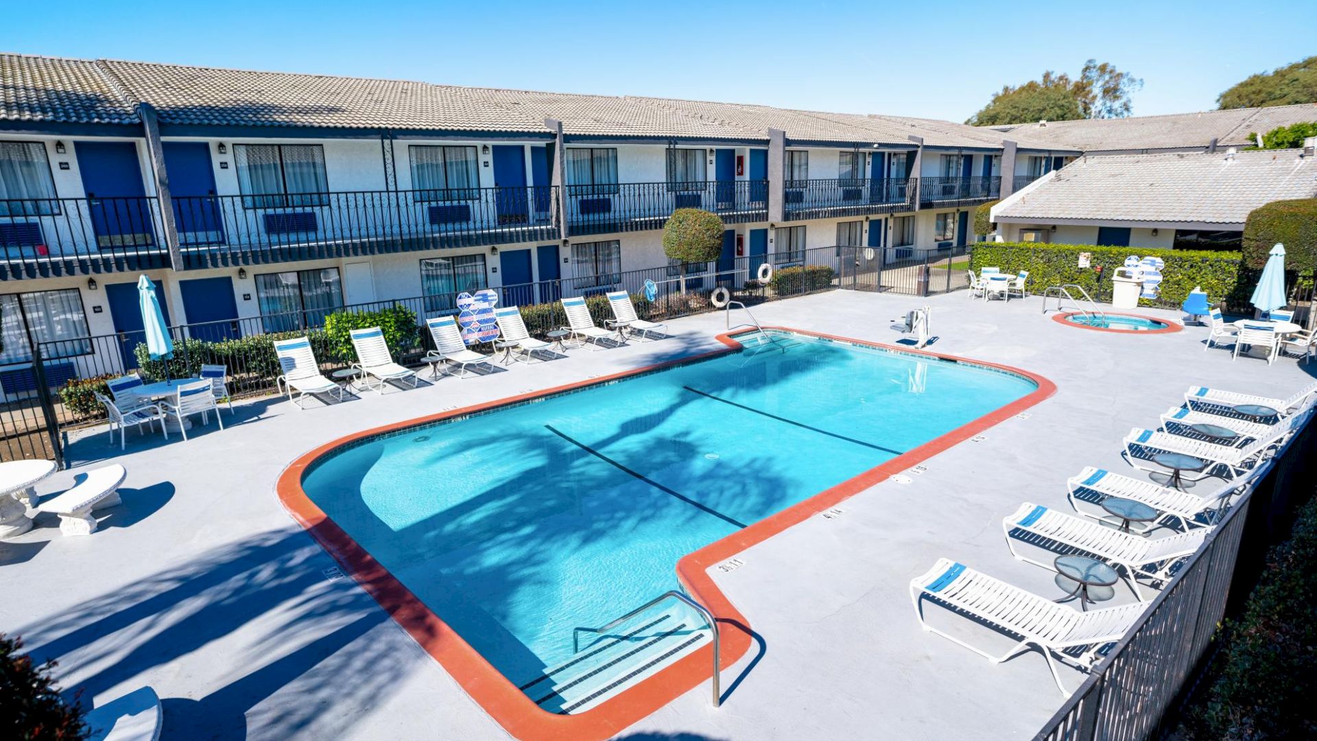 A hotel courtyard with a swimming pool, lounge chairs, and two-story buildings in the background, surrounded by a clean, paved area.
