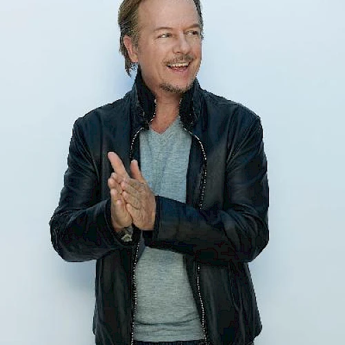 A man with blonde hair is clapping and smiling, wearing a black leather jacket over a gray shirt and blue jeans, against a plain background.