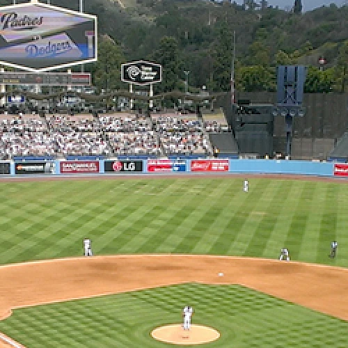 A baseball game in a stadium with players on the field and many spectators in the stands. Billboards and a scoreboard are visible in the background.