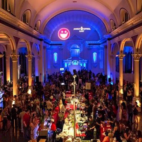 This image shows a crowded event held in a grand, high-ceilinged hall with arched columns, featuring tables and stations, under ambient blue and purple lighting.