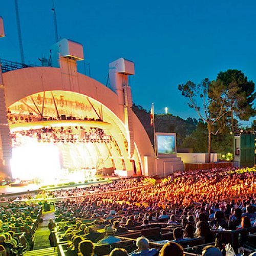 A large outdoor amphitheater is filled with a crowd watching a brightly lit stage with an archway structure under a clear evening sky.