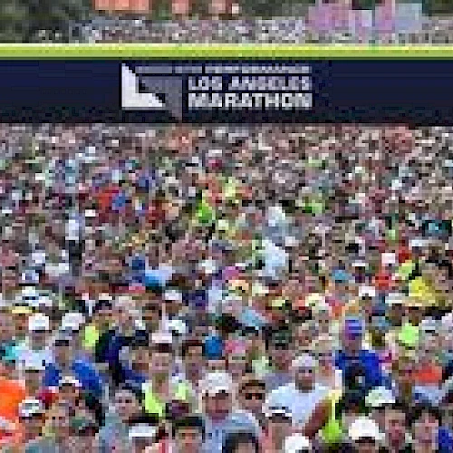 A large crowd of runners is at the starting line of the Los Angeles Marathon, with a banner overhead indicating the event.