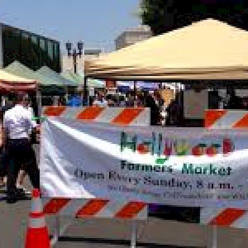 The image shows a bustling outdoor market with various colored tents and a sign that reads "Hollywood Farmers Market," open on Sundays.