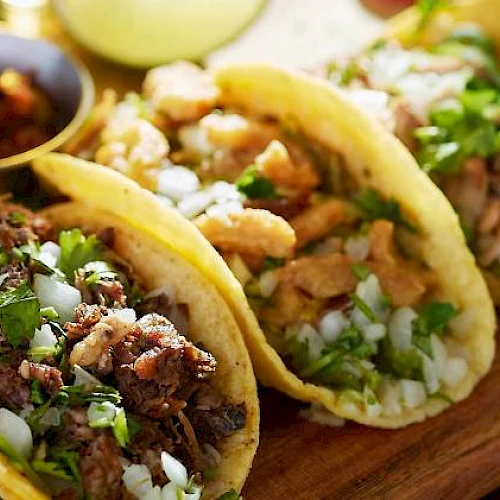 Three tacos filled with meat, cilantro, and onions are placed on a wooden board, accompanied by a small bowl of salsa in the background.
