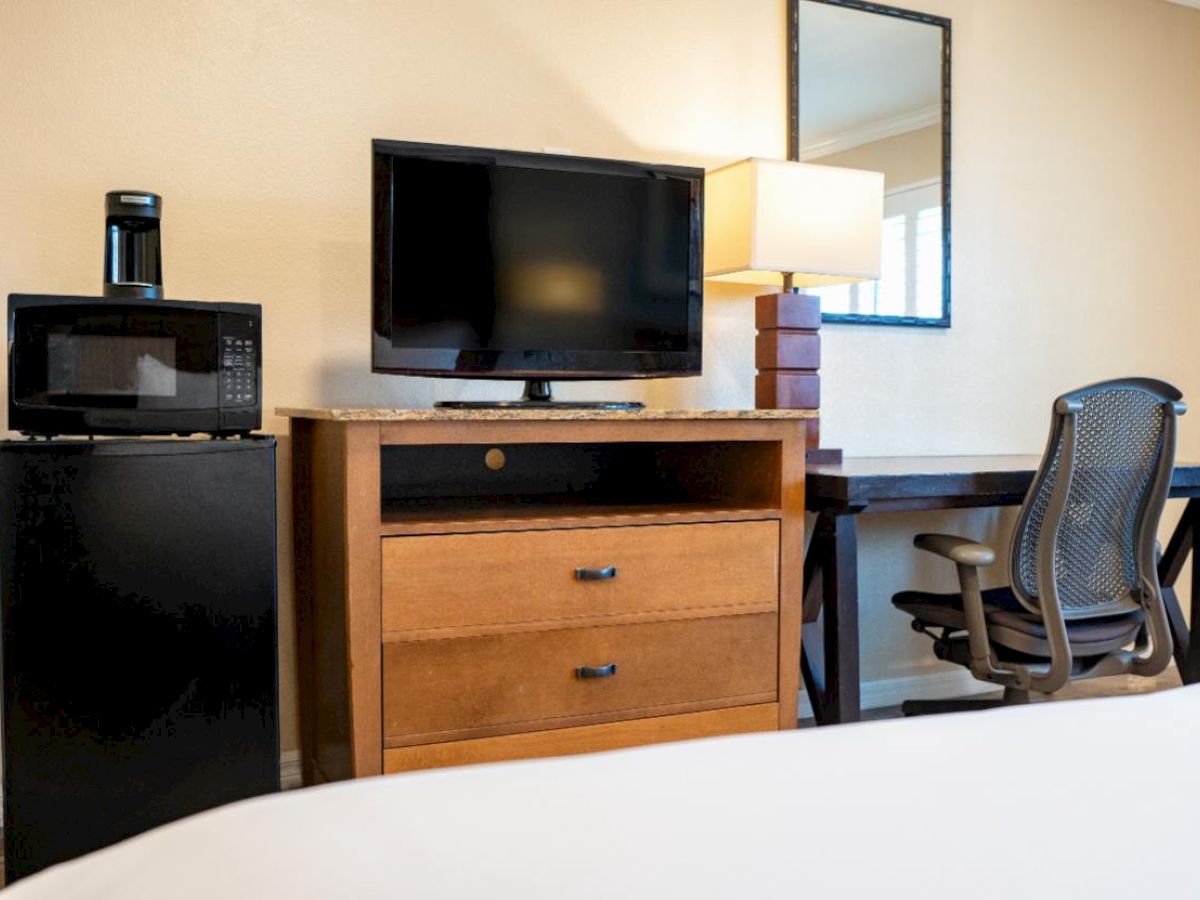 The image features a hotel room with a TV, microwave, mini-fridge, lamp, desk, chair, and mirror.