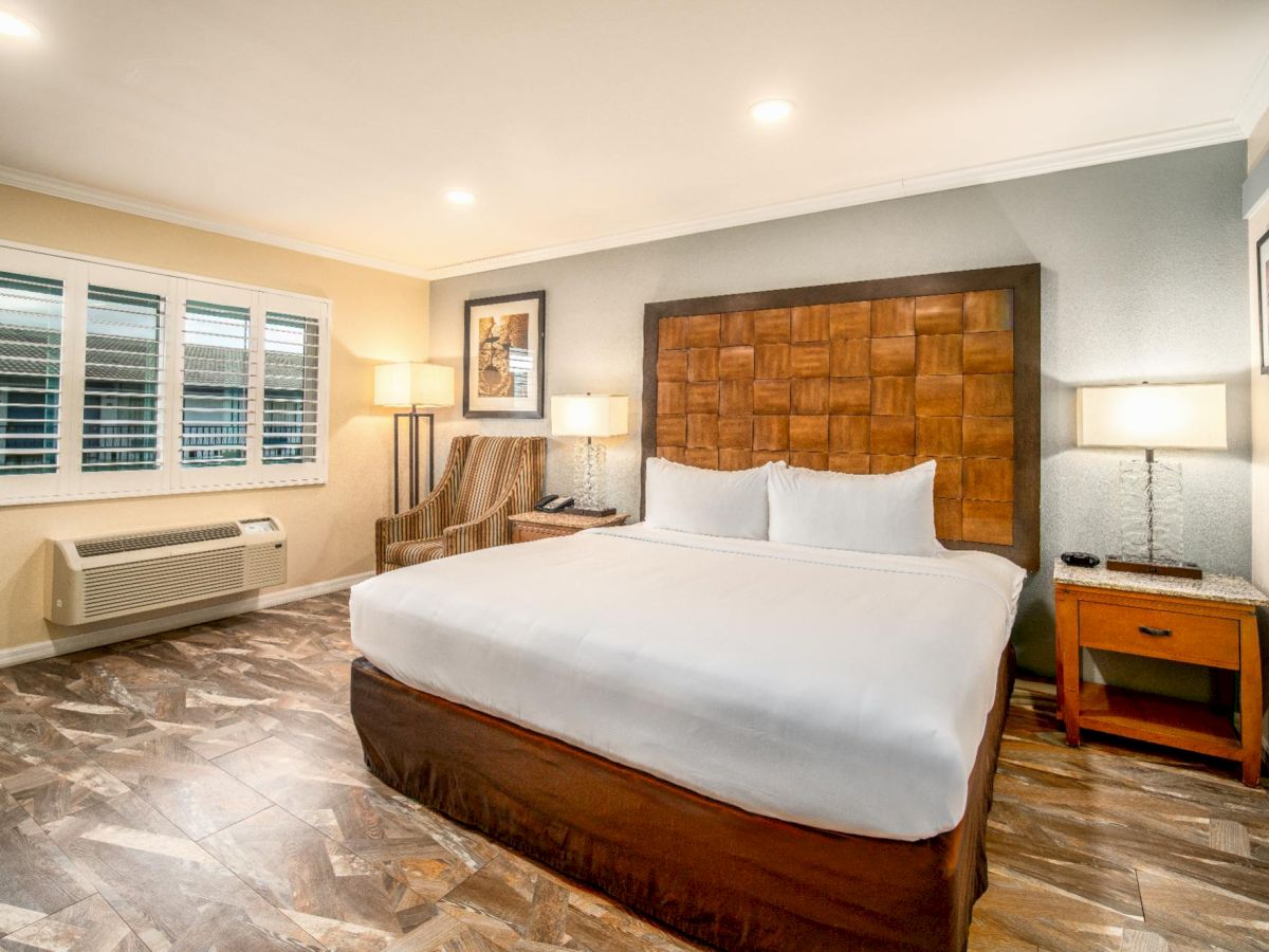 The image shows a well-lit hotel room with a large bed, two nightstands, lamps, a chair, framed paintings, and light brown tiled flooring.