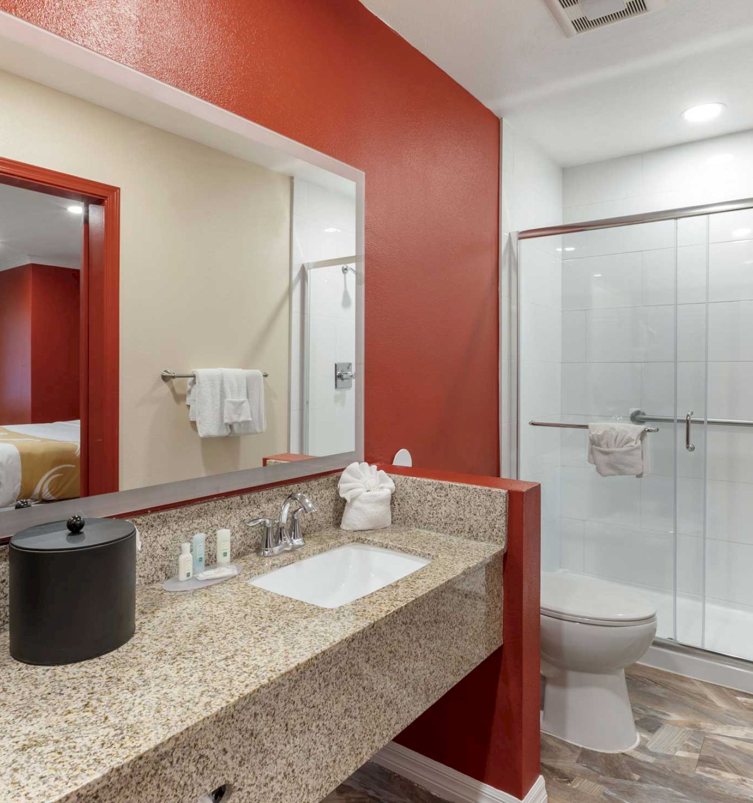 A bathroom with a granite countertop, sink, toiletries, coffee maker, mirror, and a glass shower; a bed is visible through a doorway.