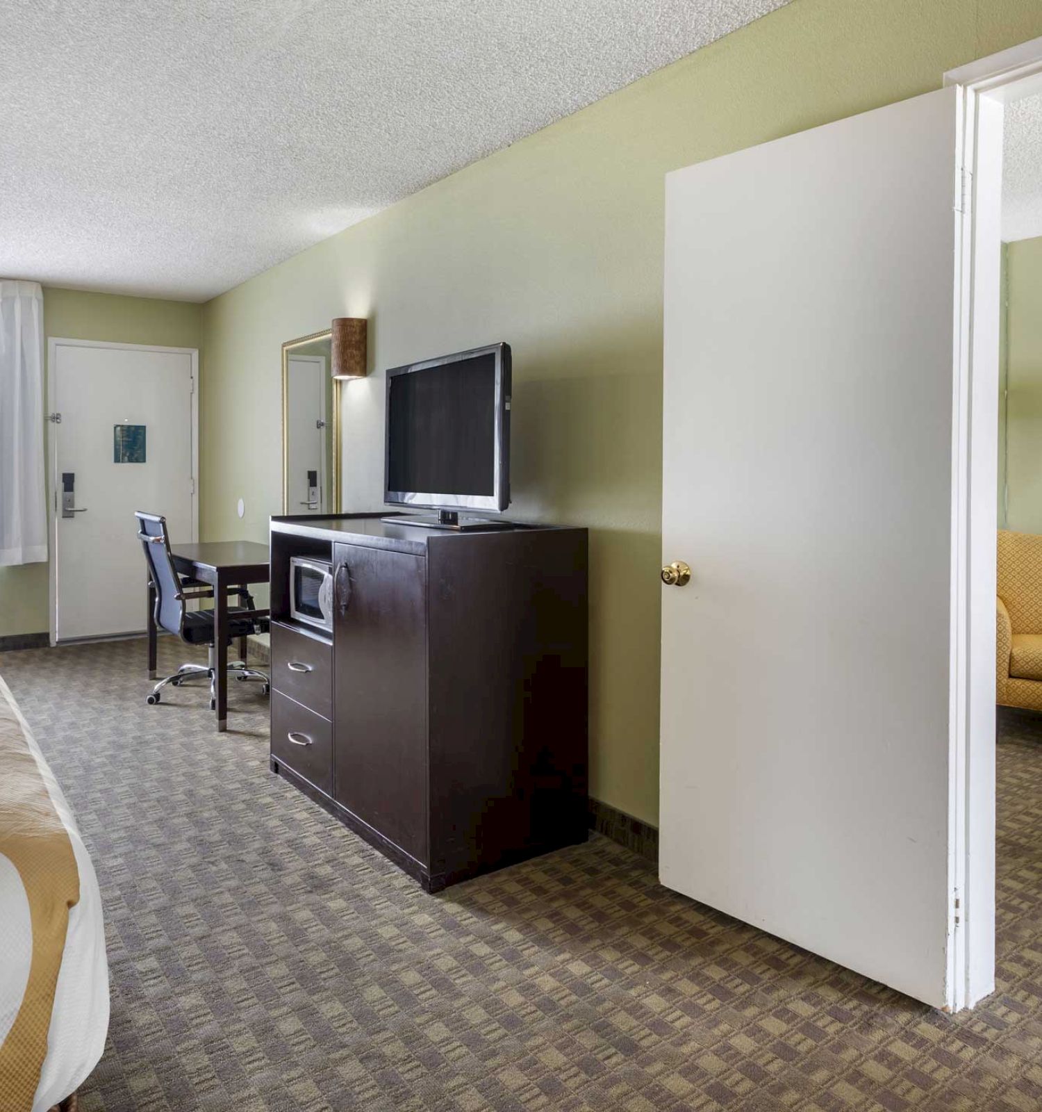 The image shows a hotel room with a bed, a dresser with a TV, microwave, desk, chair, and an adjacent room with a bed and an armchair.