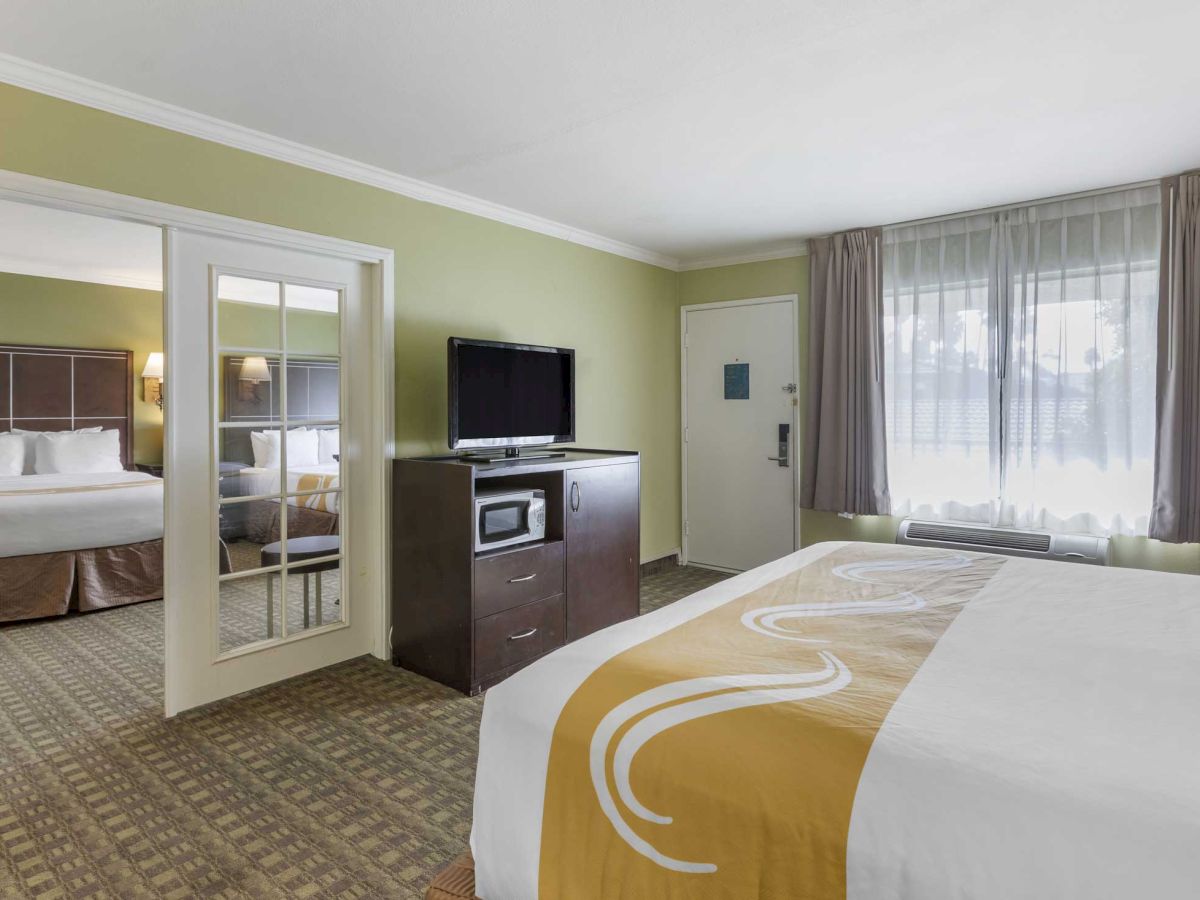 A spacious hotel room with a bed, TV, microwave, and a connecting doorway to another room with a bed. Sunlight filters through the window curtains.