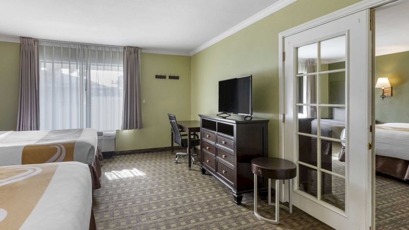A hotel room with two beds, a desk, a dresser with a TV on top, and a closet with a mirrored door.