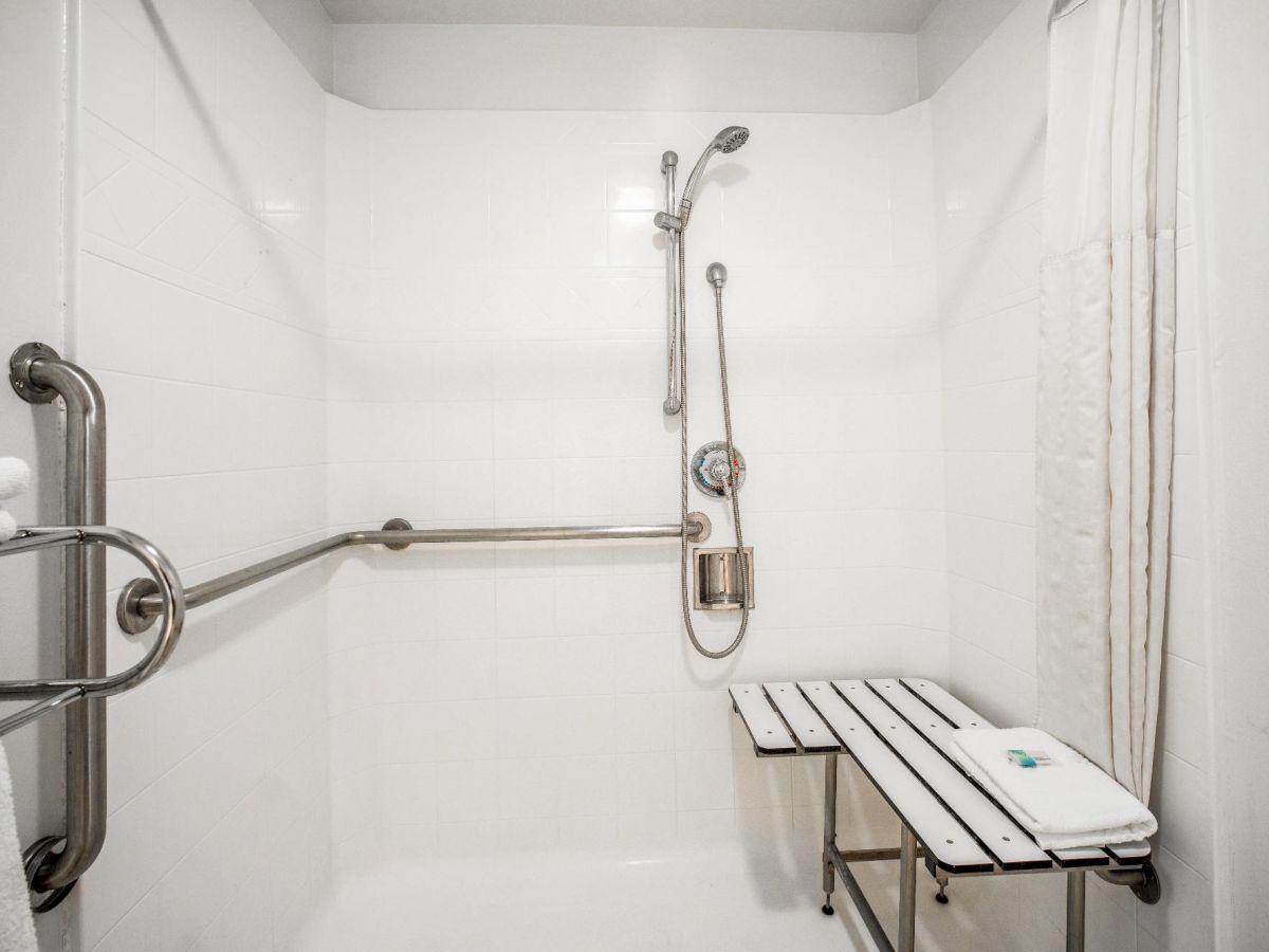 The image shows a white-tiled accessible shower with a wall-mounted bench, grab bars, a handheld showerhead, and rolled-up towels on the side.