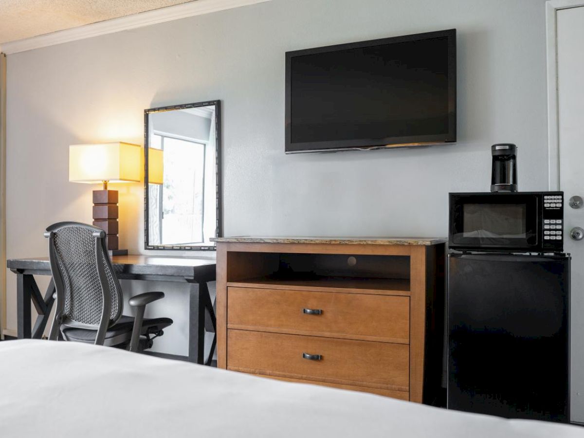 A modern hotel room features a TV, desk, chair, lamp, mirror, dresser, microwave, mini-fridge, and coffee maker.