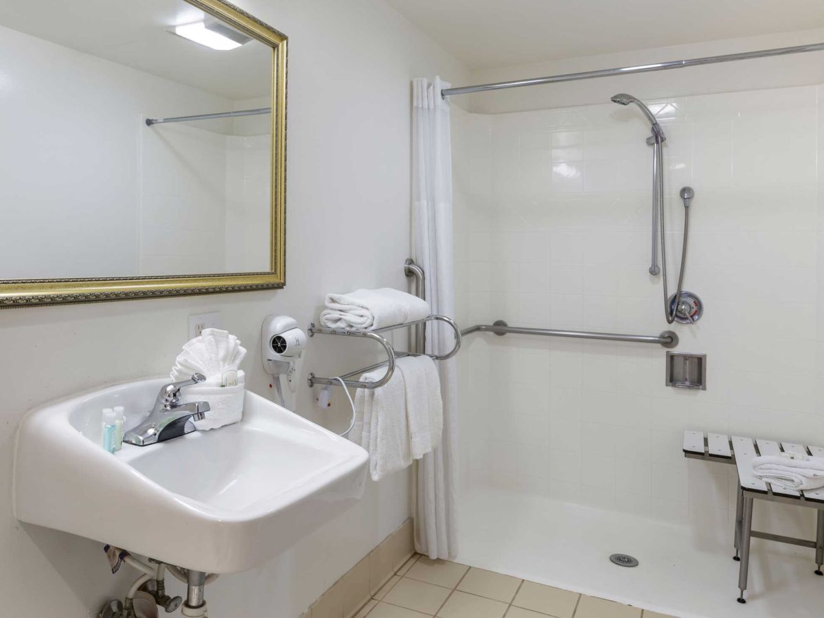 A bathroom with a sink, mirror, shower with safety bars, and a folding chair. Towels are neatly arranged near the sink and on a rail.