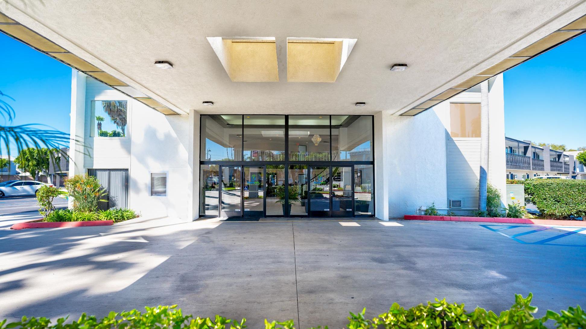 A white building with a glass entrance and an awning, surrounded by plants and a parking lot.