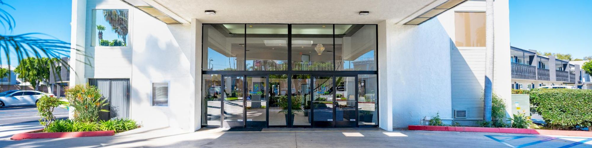 The image shows the entrance to a building with glass doors and a covered driveway. There's a well-maintained landscape with shrubs in the foreground.