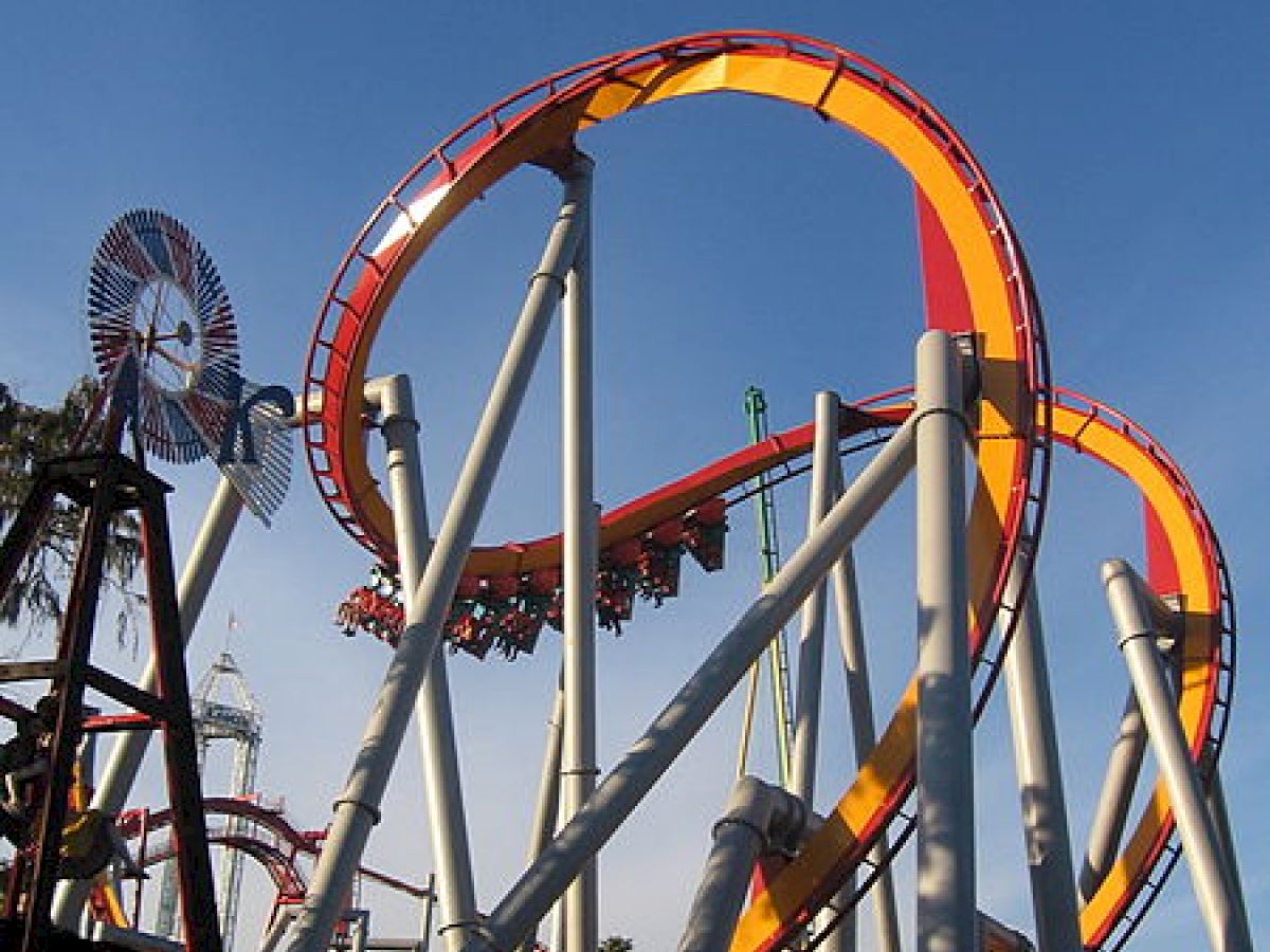 The image shows a roller coaster with orange and red tracks featuring several loops and tall supporting structures under a clear blue sky.