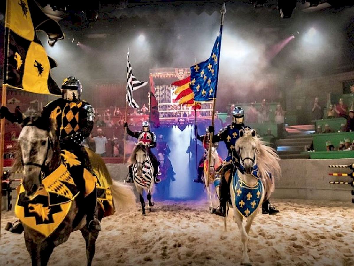 Knights on horseback, adorned in colorful medieval attire, carrying flags in an arena with an audience in the background.