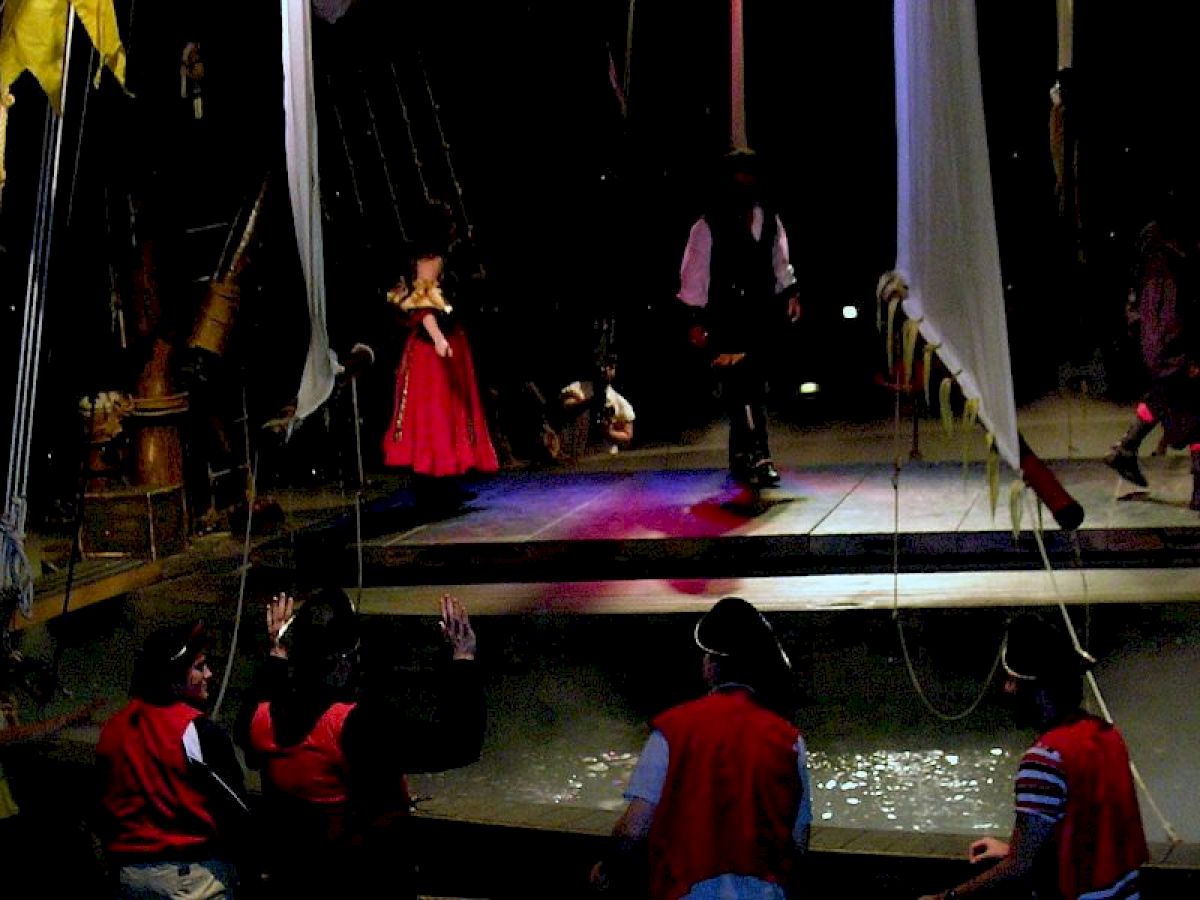 The image shows a stage scene resembling a pirate ship, with people dressed in pirate-themed costumes and one person in a red dress.