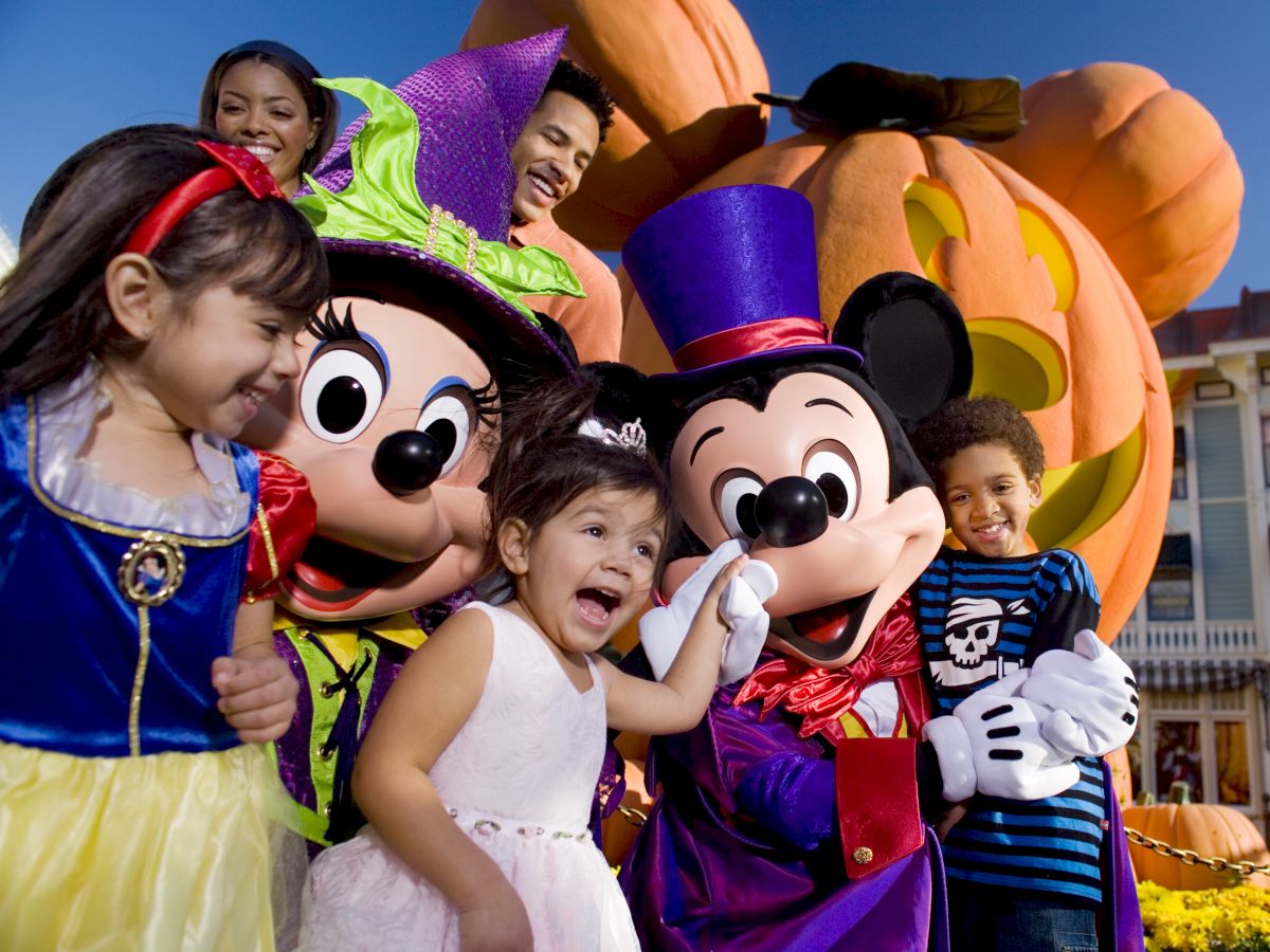 A group of children and adults are posing with costumed characters, including Mickey Mouse and Minnie Mouse, in front of large pumpkins in a festive setting.