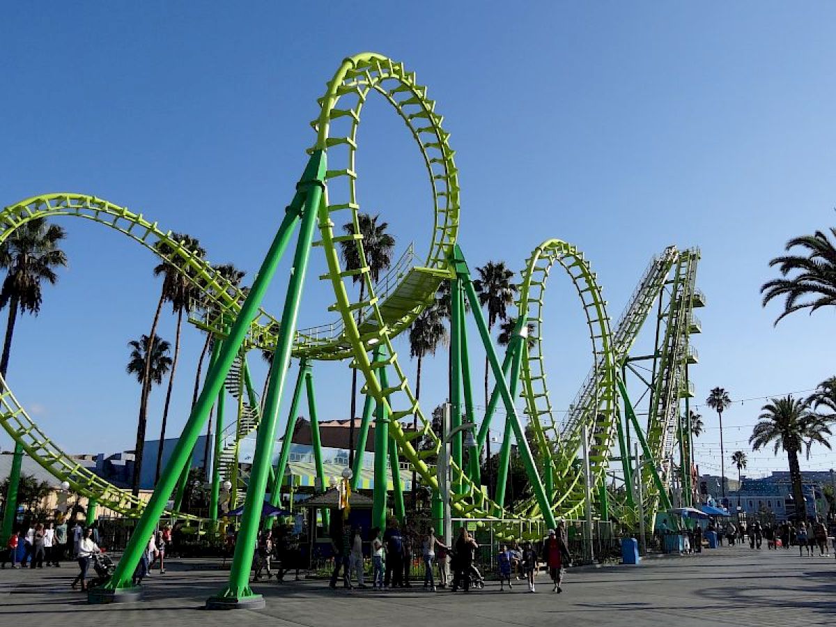 The image shows a vibrant green roller coaster at an amusement park, with palm trees and a clear blue sky in the background.