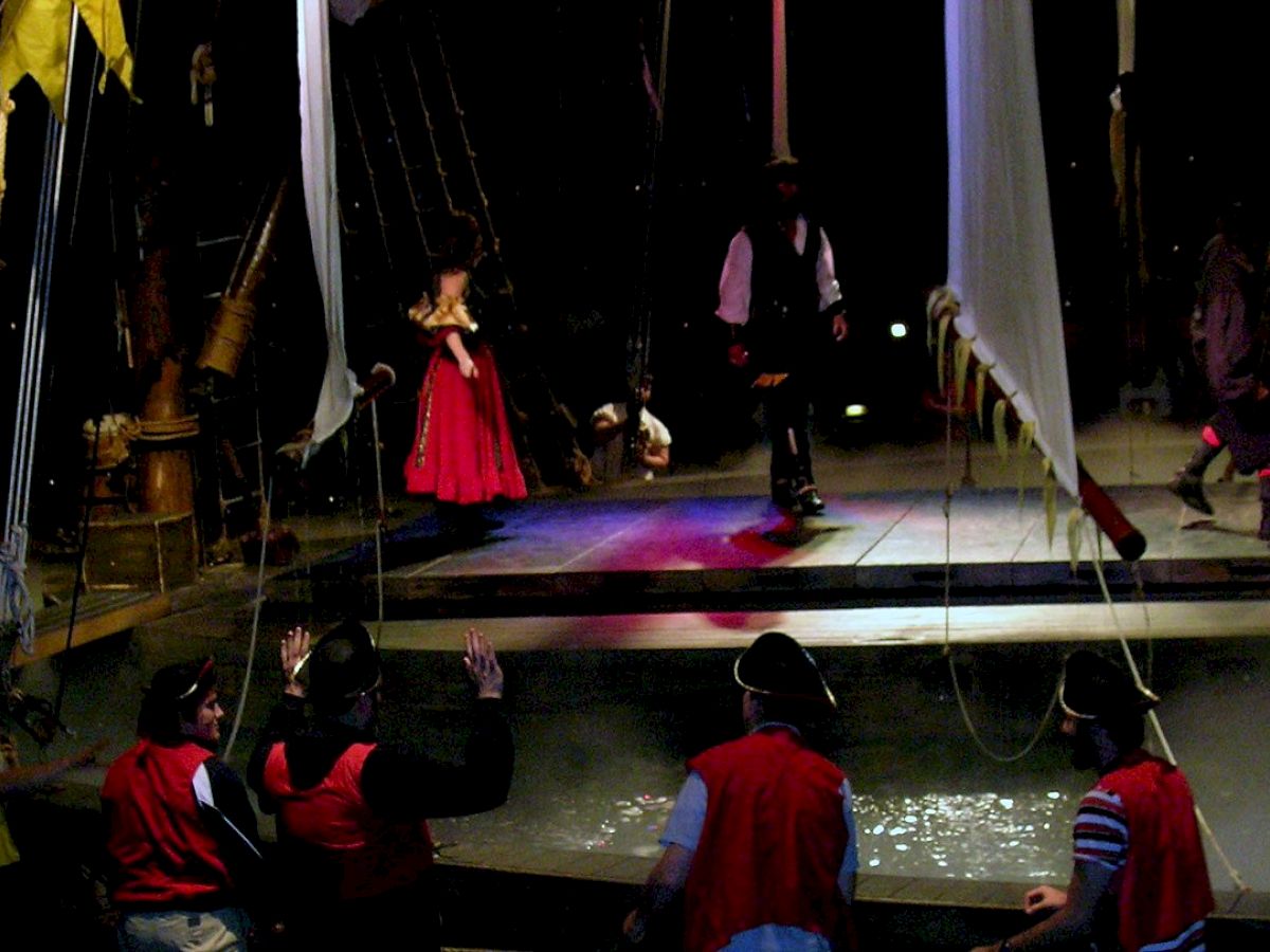 People dressed as pirates are performing on a ship-themed stage with a crew and audience engaging in the scene, creating a lively atmosphere.