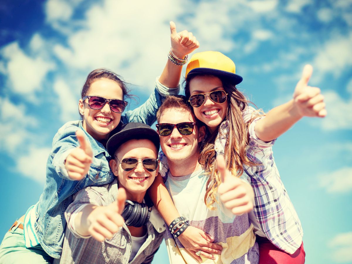 Four friends wearing sunglasses and casual clothes are smiling and giving thumbs up under a bright blue sky with scattered clouds, looking happy.