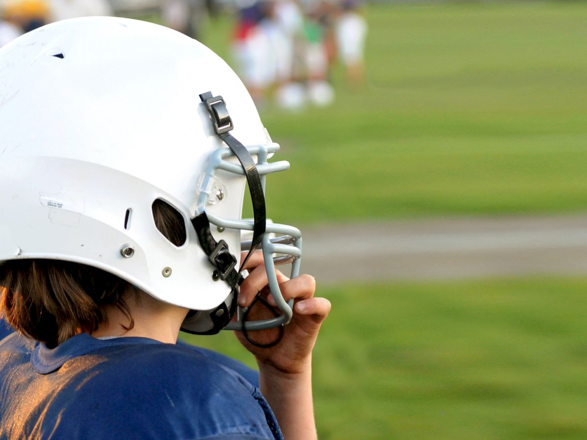 A person wearing a white football helmet and blue shirt looks out at a grassy field, with blurred figures in the background.