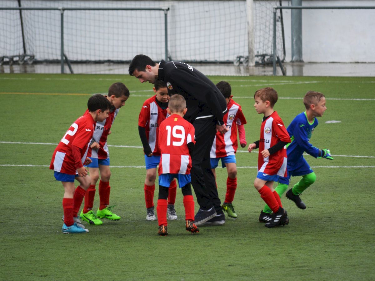 A group of children in red and white soccer uniforms are huddled around a coach on the field, receiving instructions before a game.