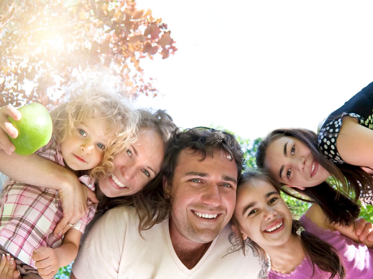 A family of five smiles while huddling together outdoors, with the child on the left holding a green apple.